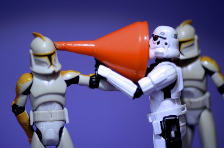 A tory robot holds a funnel up to another robot as if using it to listen to them.