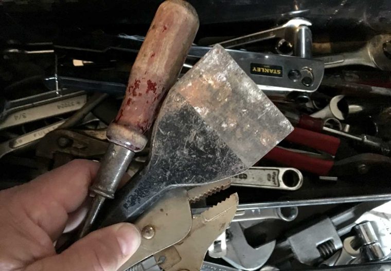 An old screwdriver, chisel, and pliers in a hand over a box of many more tools