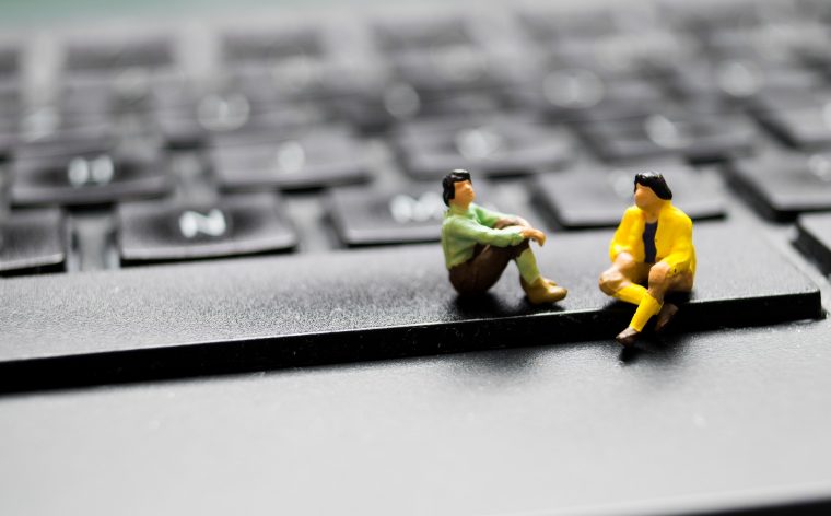 Two tiny toy human figures sit as if in conversation on a giant computer keyboard