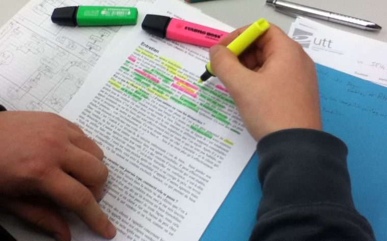 A hand holds a yellow highlighter pen over a document with pink and green highlights (these pens can be seen on the side)