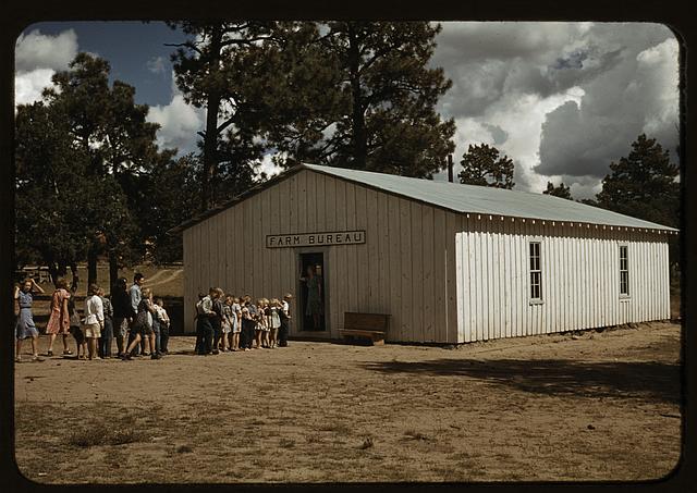 A line of kids and a few adults, dressed as if in the times of the 1940s, are lined up to enter a meta shed structure bearing the label "Farm Bureau"