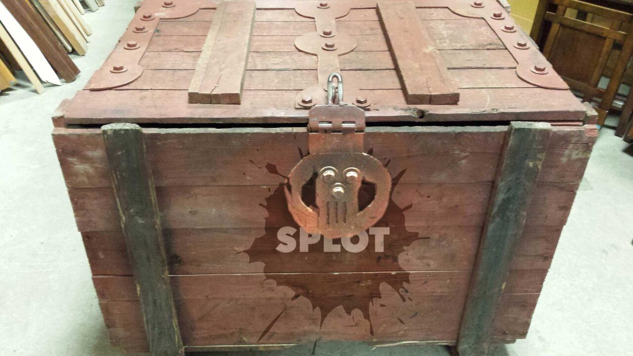 A large wooden box, old style like it was made by hand, with heavy metal braces and a clasp. Superimposed on front is a pattern of a paint blob with the word SPLOT in the middle.