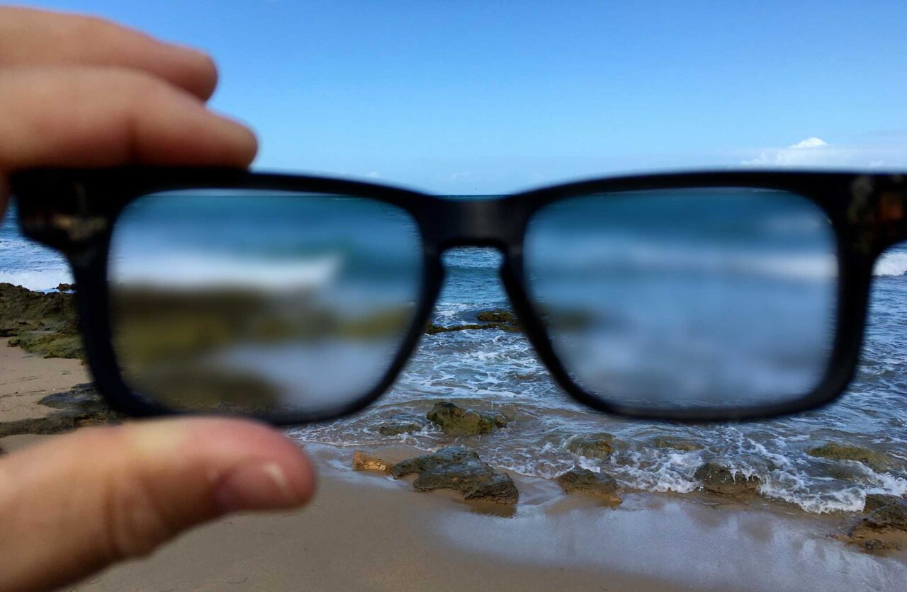 A hand on the left holds a pair of eye glasses looking at a view of a beach, but the view through the glasses is blurred