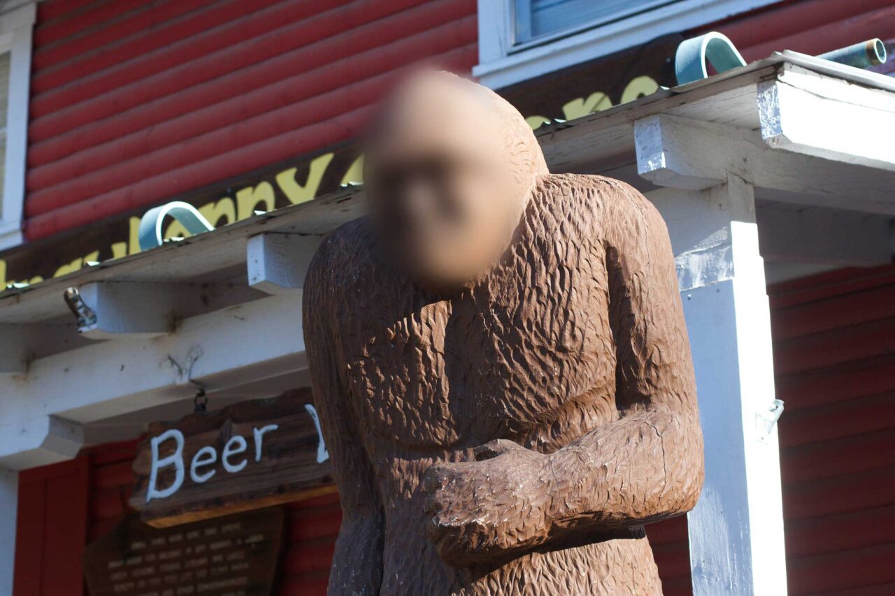 A statue of a large brown creature, it's face blurred, in front of a red, wooden front store