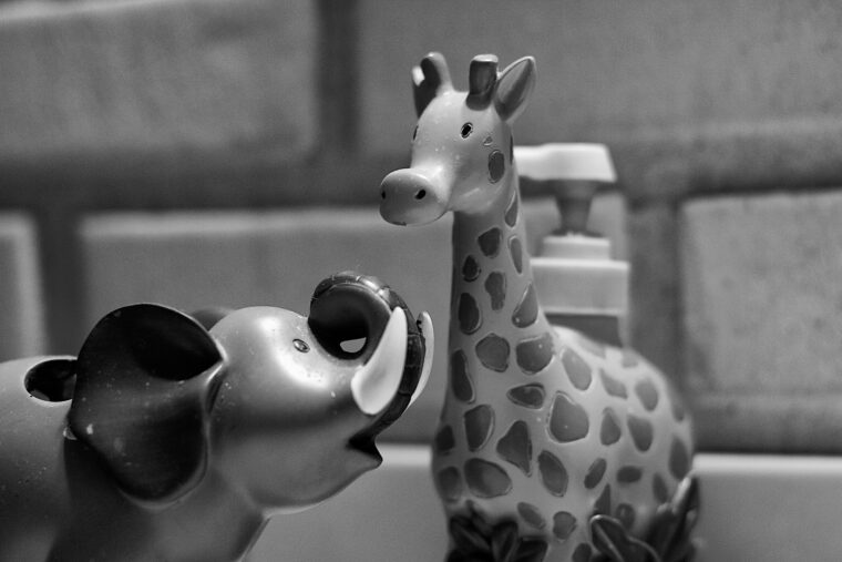Black and white photo up close to a toy elephant on left looking up to a taller toy giraffe, in front of a faux brick wall