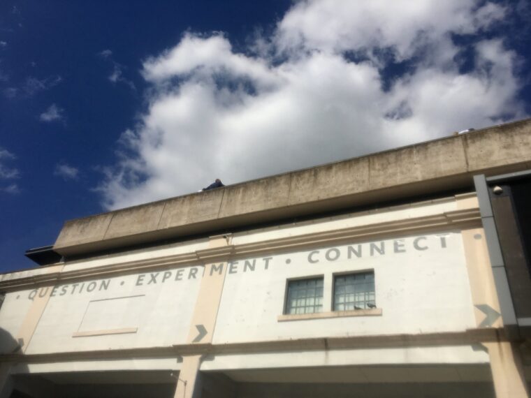 Looking upward at a 1970s style building, plain walls, just two windows with birds in front. Words at the top of the building trim read Question - Experiment - Connect