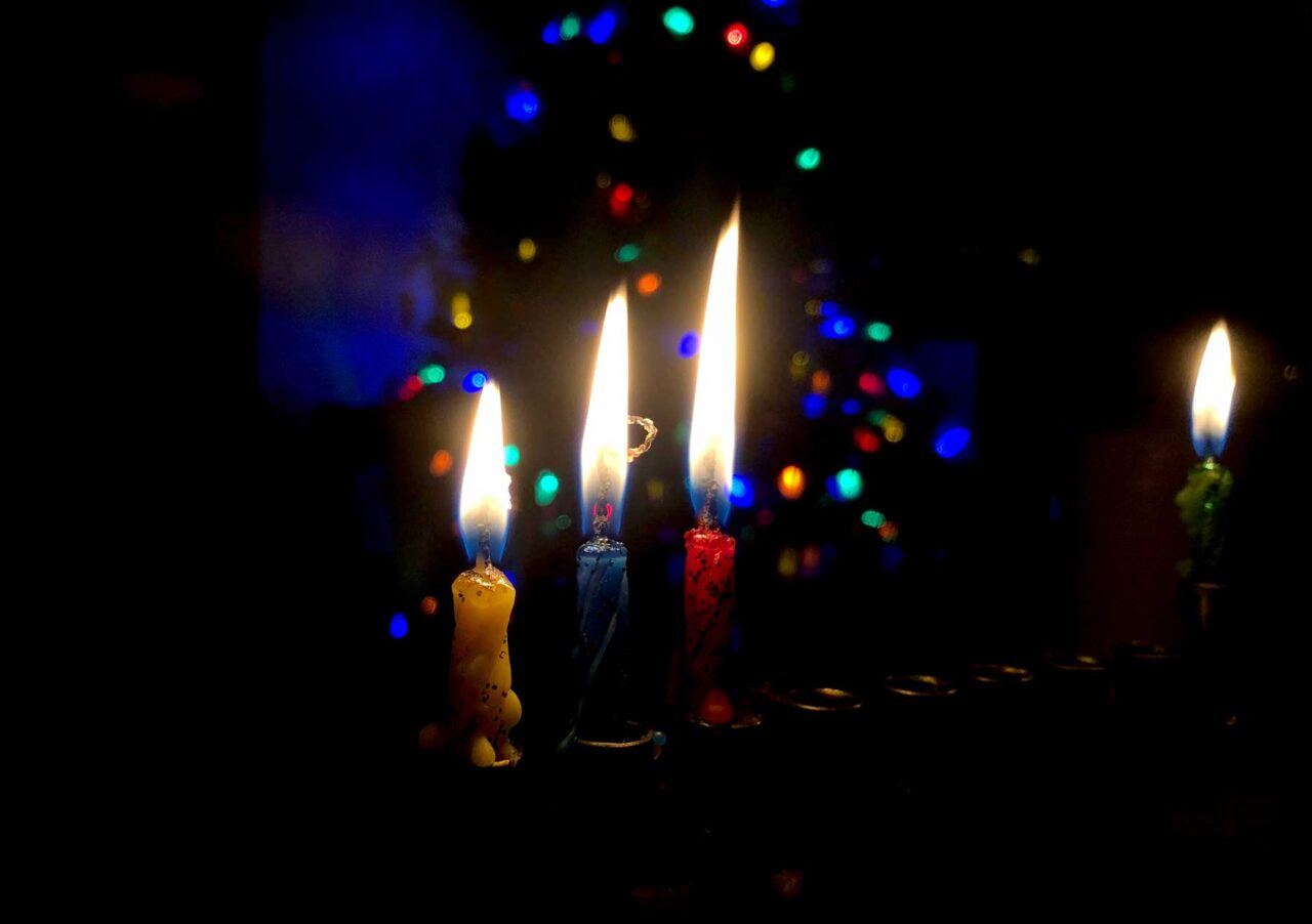 Three Hanukkah candles lit in front of lights from Christmas Tree.