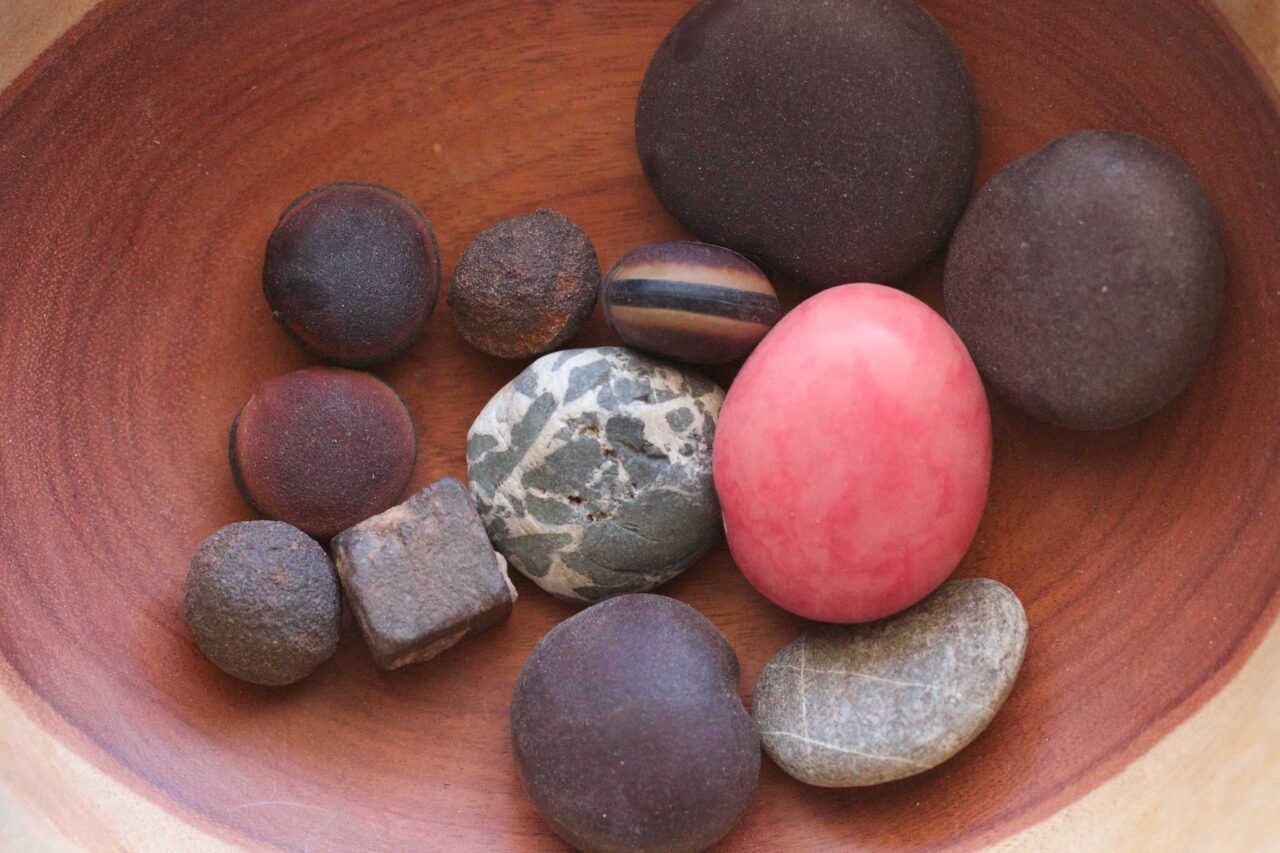 A wooden bowl full of various round stones, most dark, but one shiny pink, another is square, and one even looks like a hamburger.