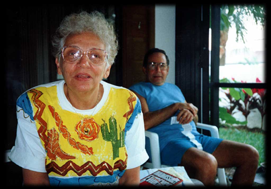 My silver haired mom leaning close to camera, sporting a colorful cactus themed short, my dad further back in his blue shirt and shorts, sitting together in shade of their Florida porch