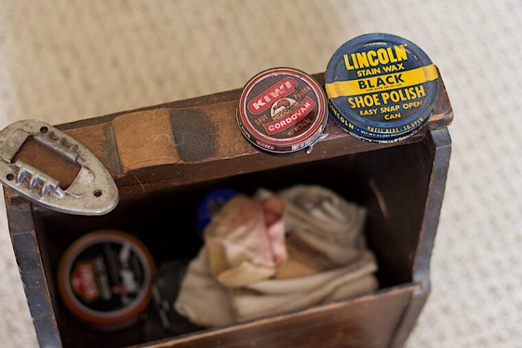 A home built thin wood carrying box with rags and cans insider, and on top are two old tins of shoe polish.