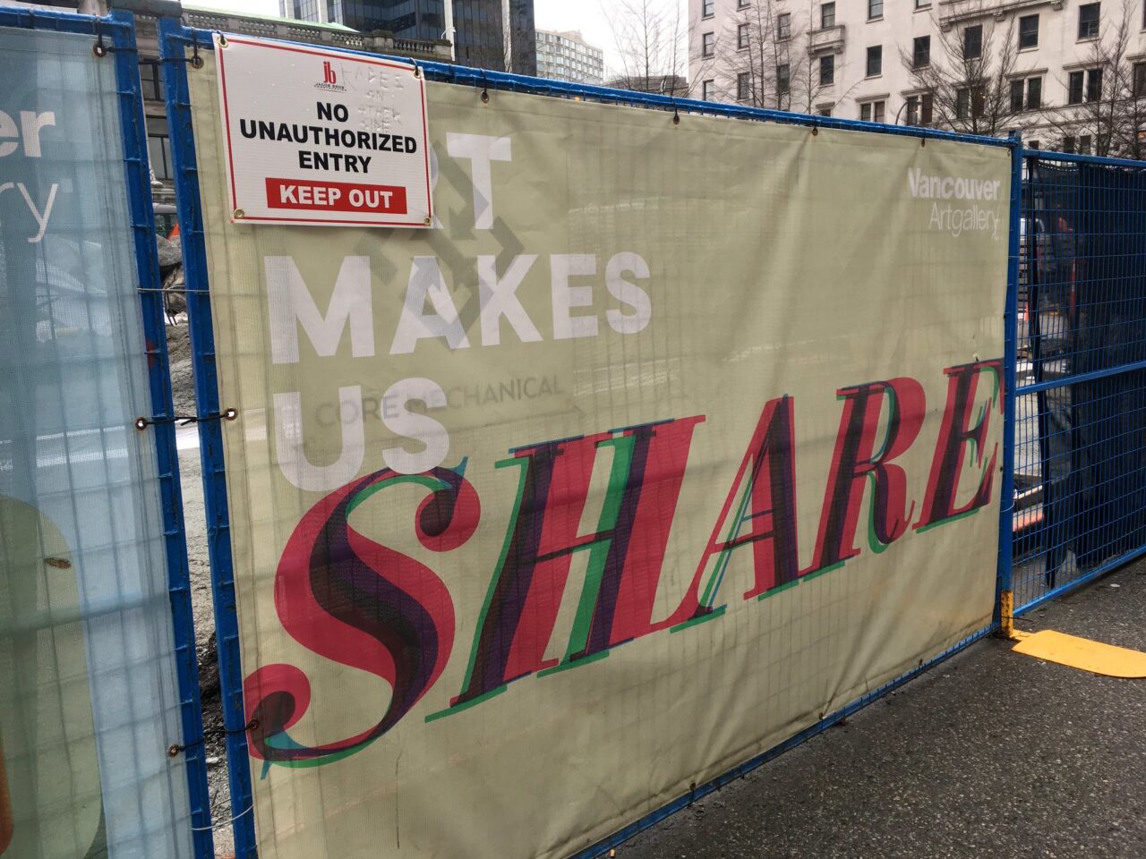 Sign on a construction fence reading in large text "It Makes Us Share" which seems in contradiction to a smaller sign that reads "no unauthorized entry keep out"