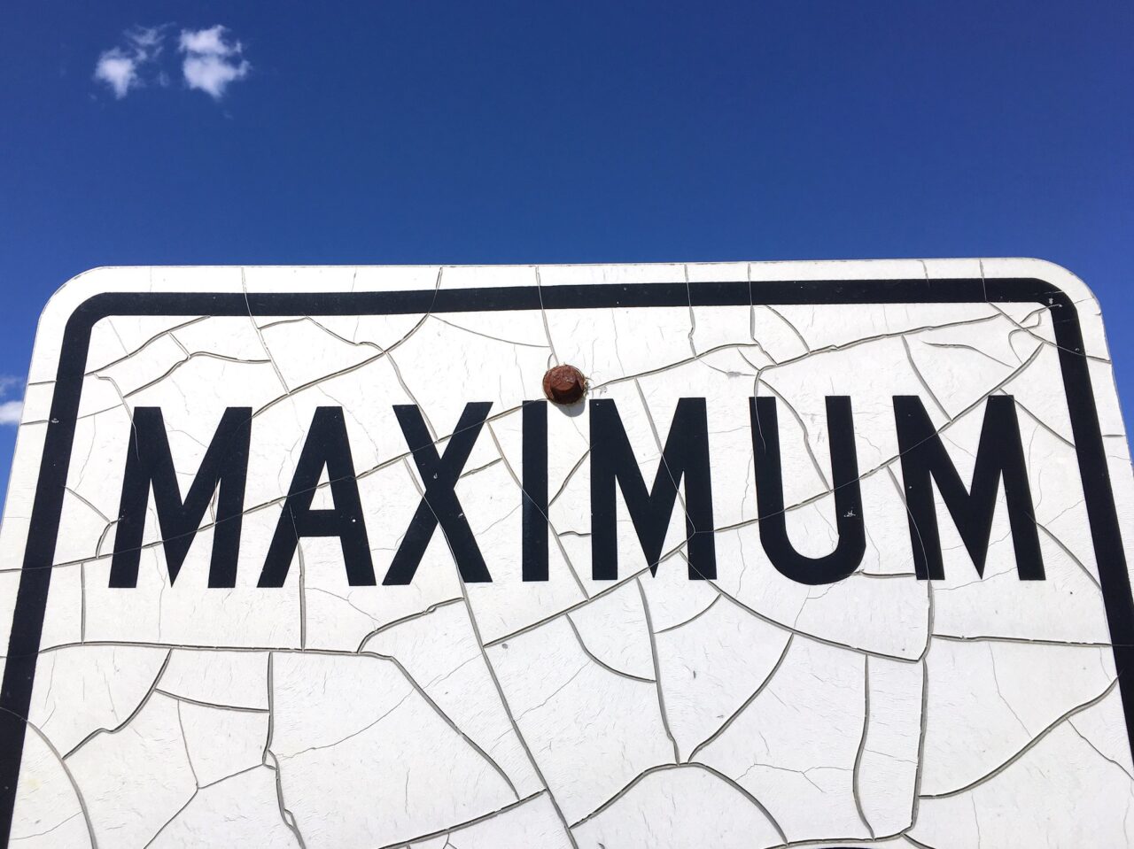 Top half of a cracked, weathered metal road sign that reads "Maximum" below a solid, cloudless sky.