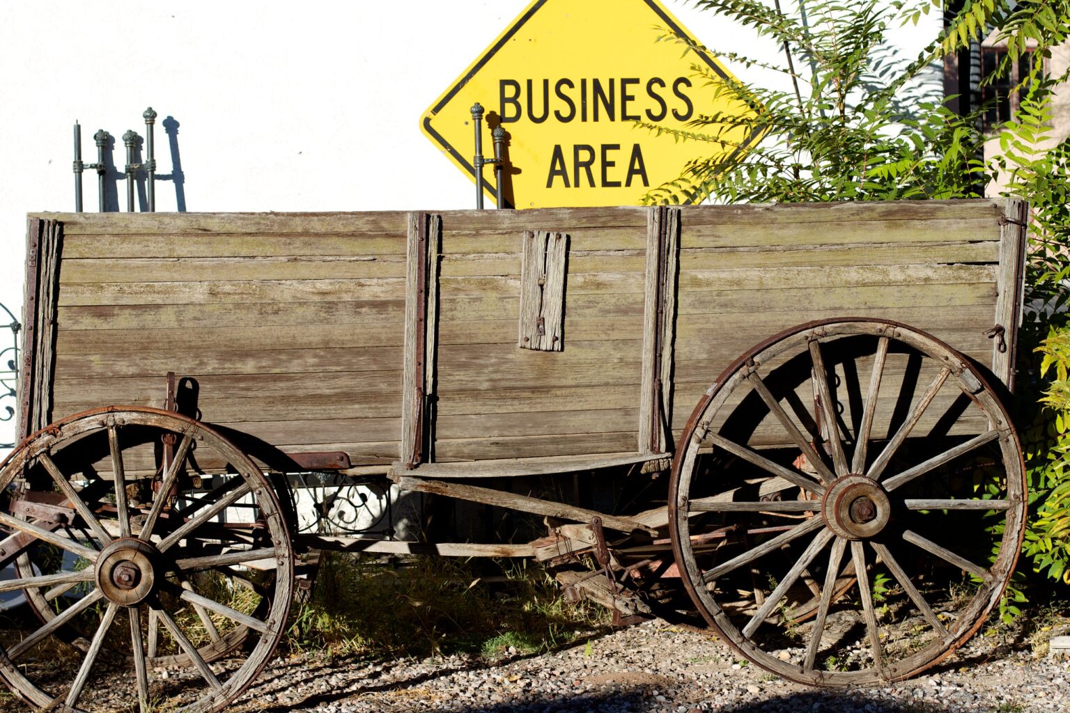 An old wooden wagon with large wheels, sitting on gravel. Behind is a modern yellow road sign that reads "Business Area"