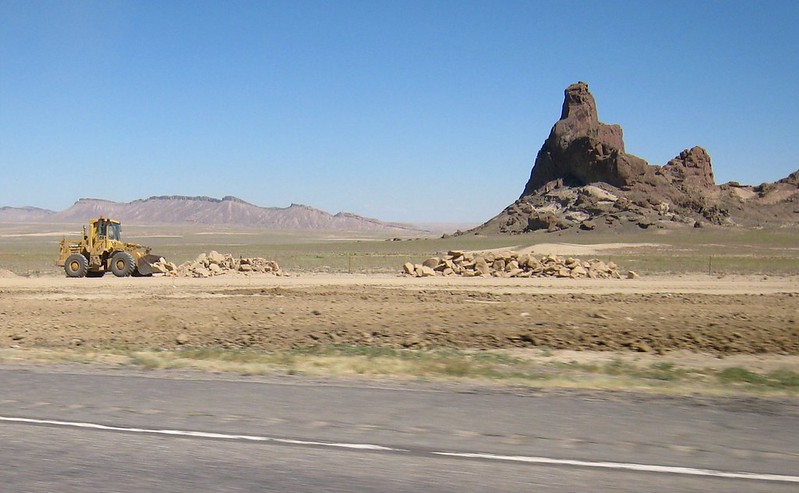 A bulldozer moves rocks beside a road in an arid landscape with a striking rock formation and distant mountains under a clear sky.