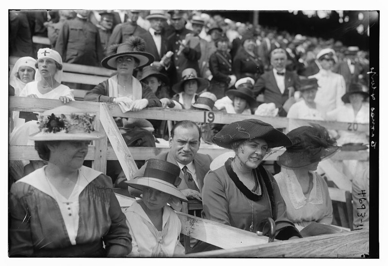 Black and white photo of a crowd at an outdoor event, with women in large hats and a man in a suit in the foreground.