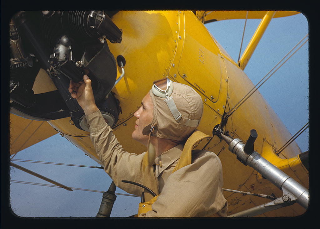 A person in a tan flight suit inspects a yellow biplane engine under a clear blue sky.