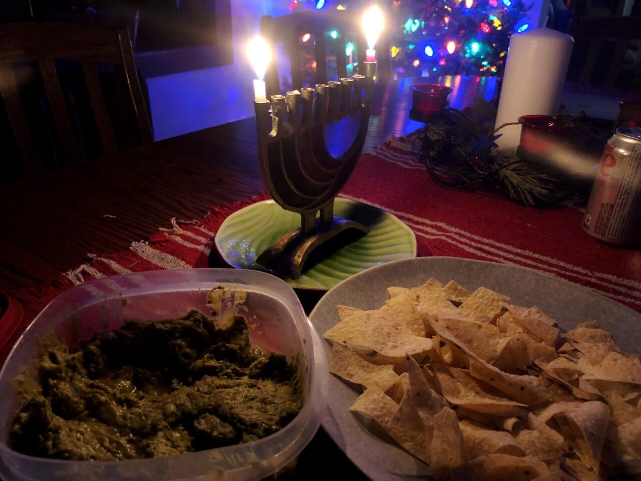 A Hanukkah memorah burns candles with a Christmas tree in te background. On the table in front is a bowl of tortilla hips and guacamole.