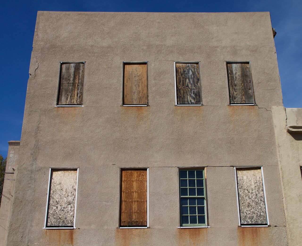Abandoned stucco 2 story building front with 8 windows, 4 on eah level, most covered in wood of various colors, ages.