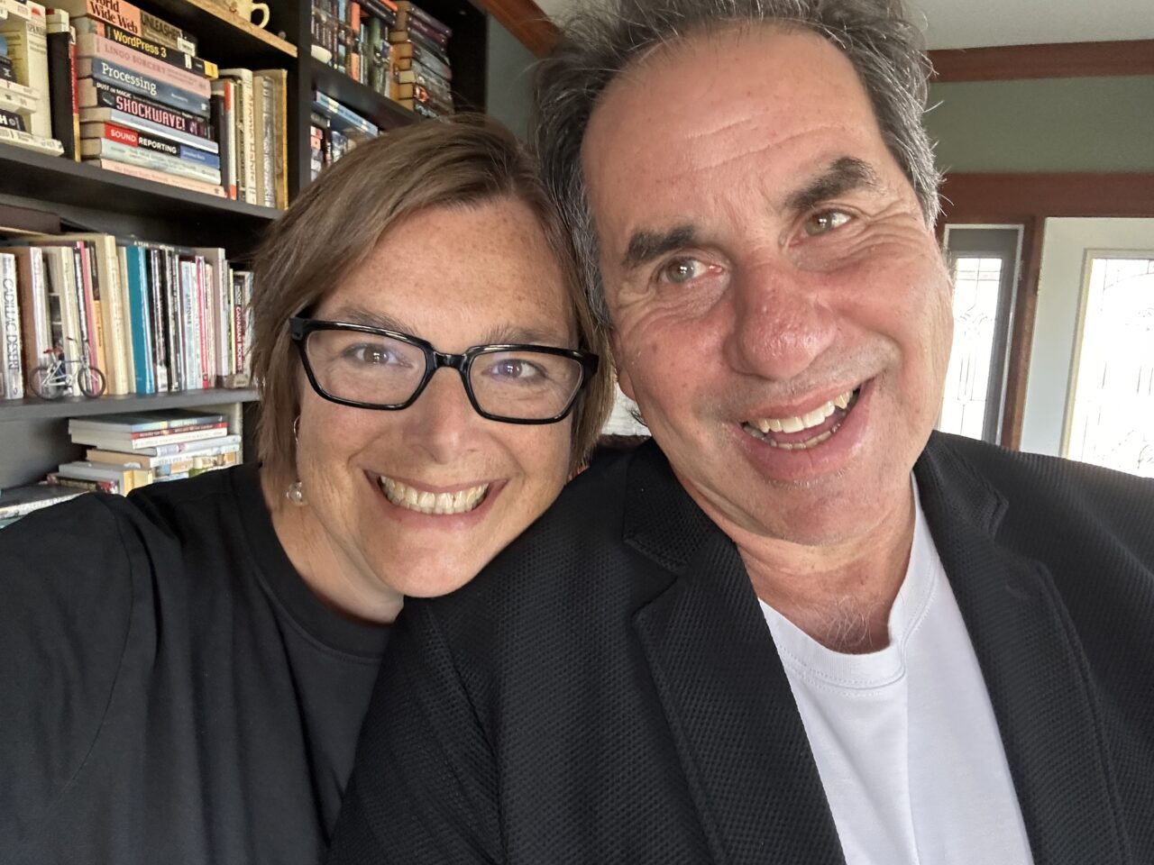 Woman in glasses and black shirt smiling broadly along side man also smiling in suit jacket, standing in front of a wall fo books. They look happy as can be.