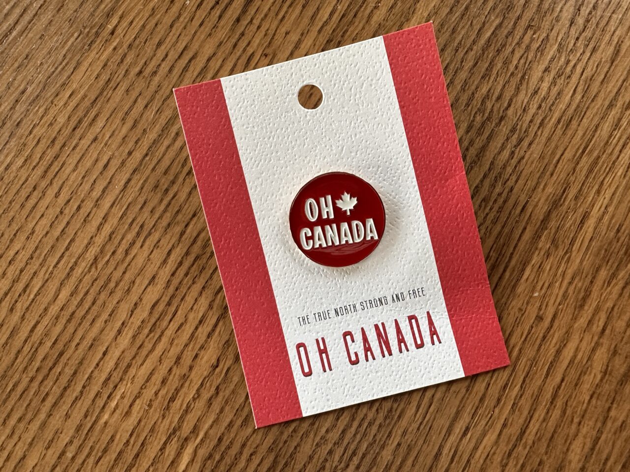 A red souvenir pin with the words Oh Canada attached to a card sitting on a wood table