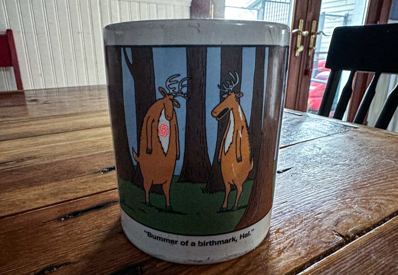 Coffee mug with Cartoon of two deer standing in a forest, one with a red OPenAI logo on its chest. The caption reads Bummer of a Birthmark, Hal.