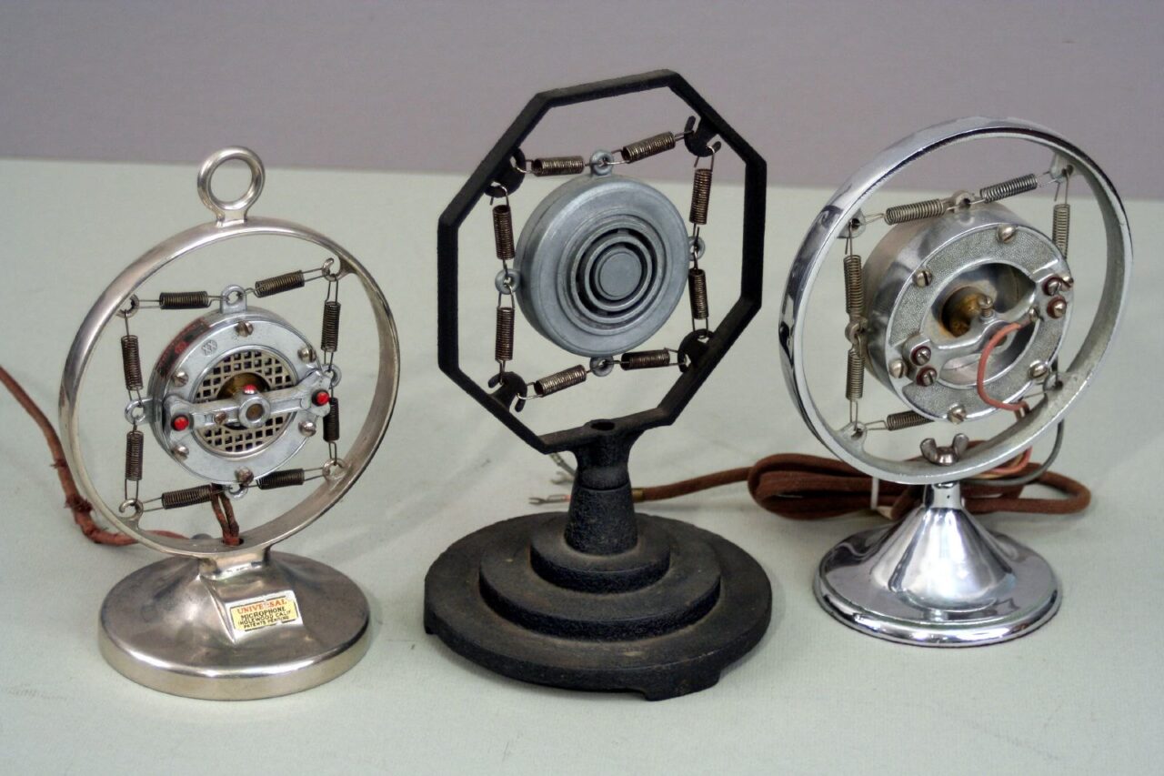 Three antique microphones sittign on a table. Each has a round frame and a suspending element in the middle.