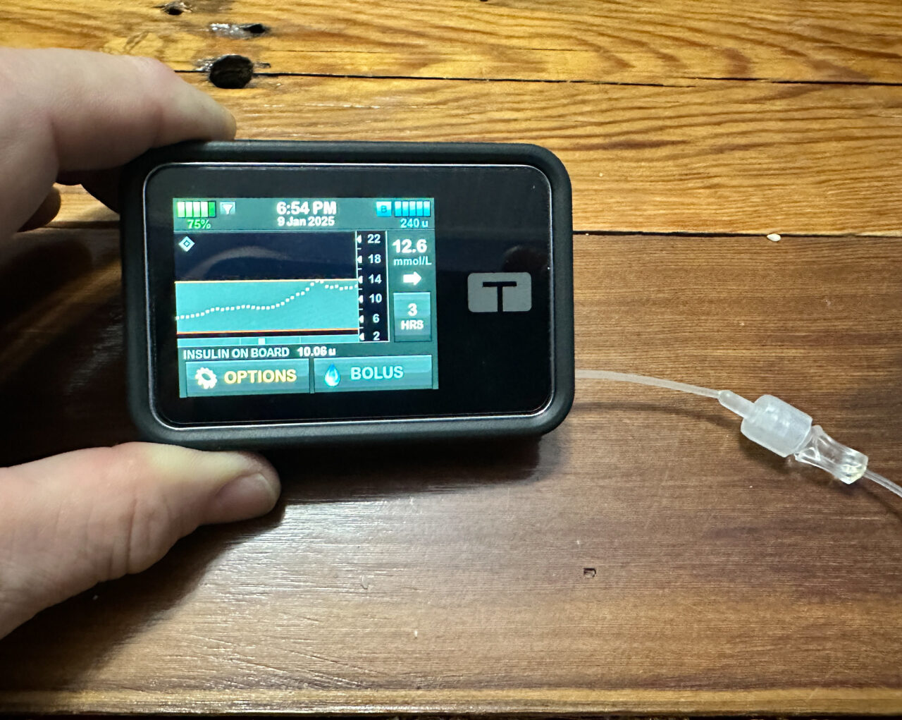 Two fingers holding an insulin pump with data display shown, sitting on a wood table.