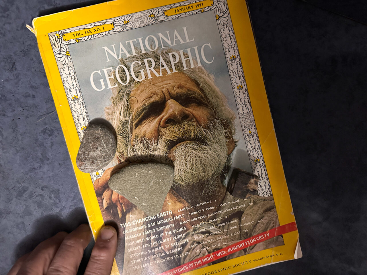 National Geographic Cover from Janaury 1973 features an old man clutching a walking stick, and the lead articles is This Changing Earth. A few rocks sit atop the magazine.