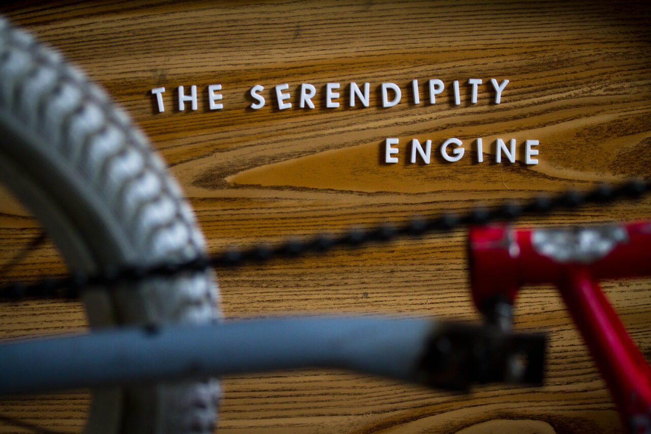 Over a wood surface, block letters placed at different angles spell out The Serendipity Machine. In the foreground and out of focus are parets of a bicycle tire, a bit of a bike frame and a chain.