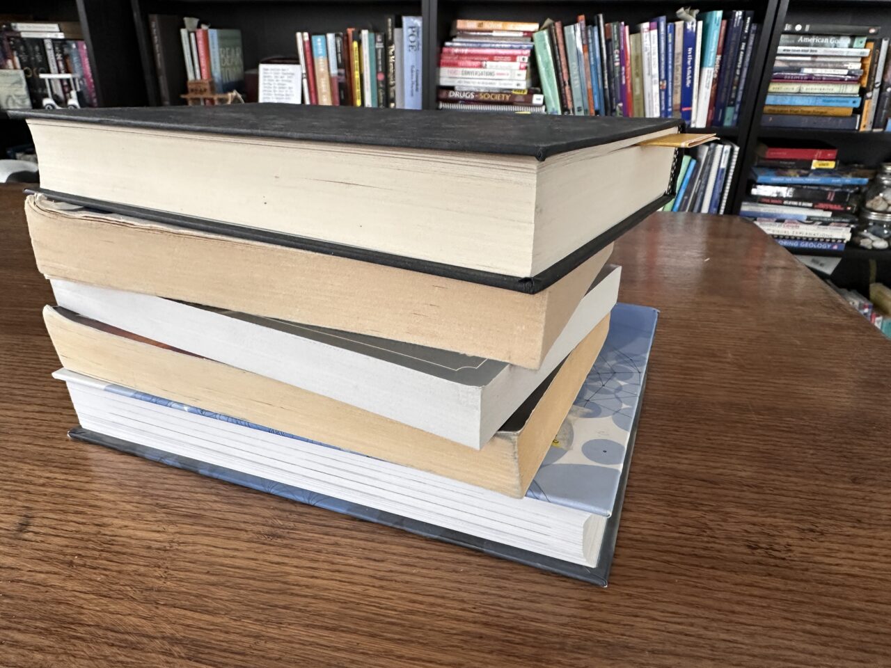 Stack of five books on a wood desk, the spines are turned to hide the titles. In the background are shelves filled with many more books.