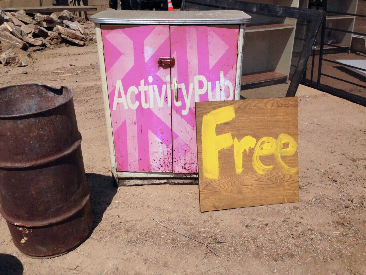 In a dirt yard is a steel barrel and a metal rusted cabinet with a hand painted sign in front reading Free. The front doors of the cabinet have been eited to include the ActivityPub plugin banner image.
