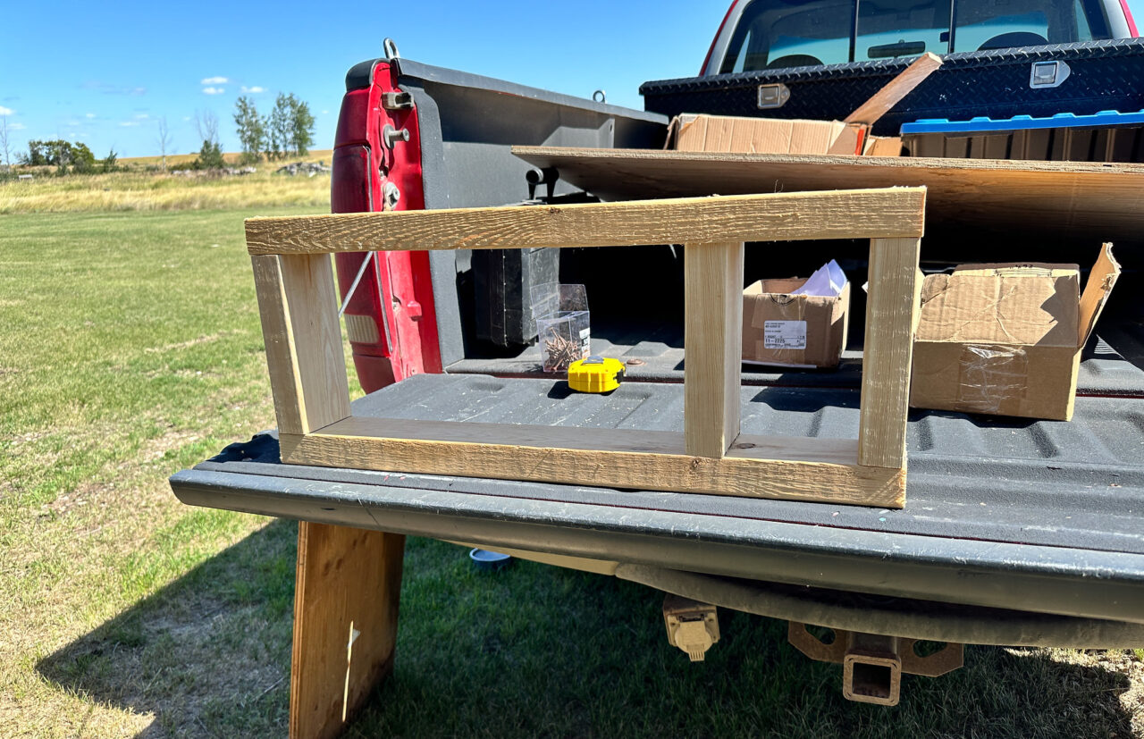 Rectangular frame of 2x4s builindg bult on ther back of a pickup truck, measuring tape and other tools, extra wood nearby.