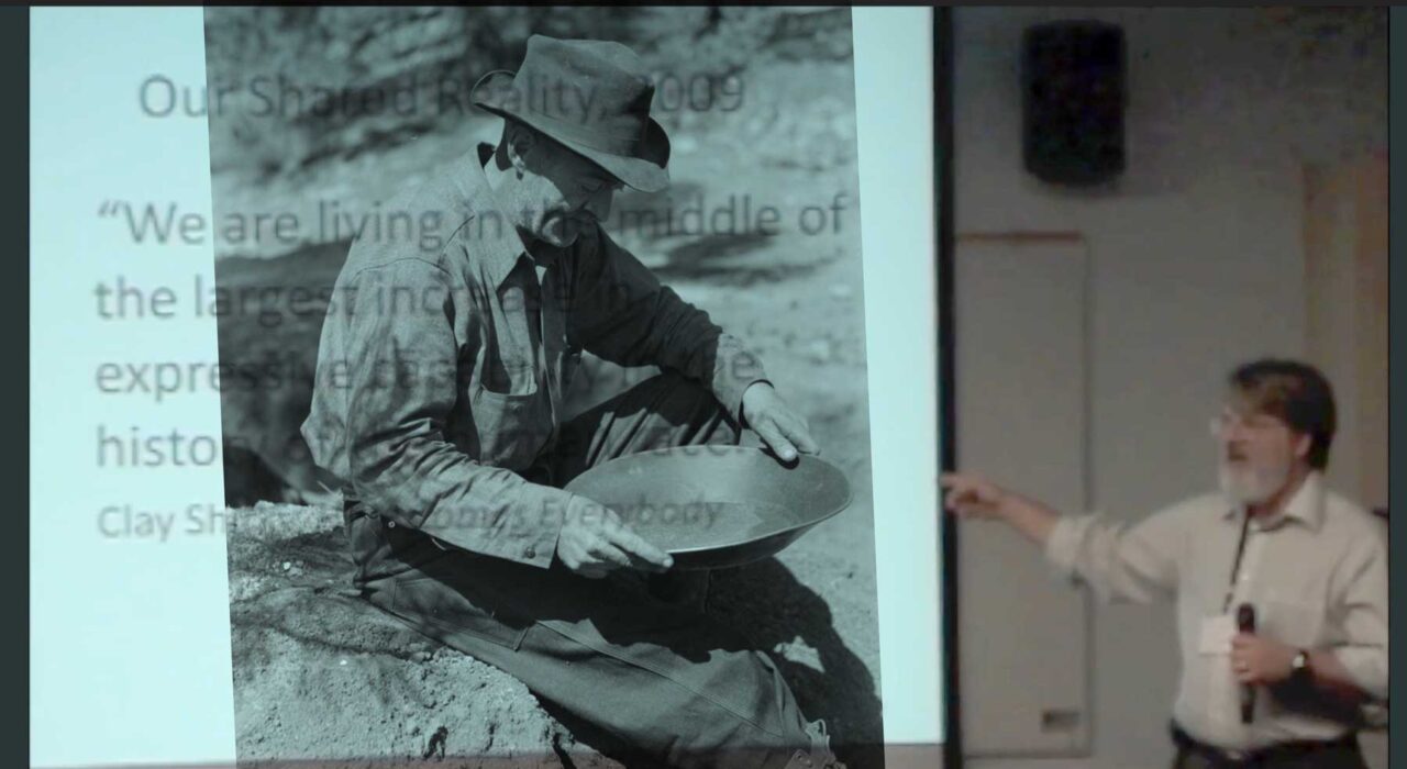 Vintage photo of a man panning for gold superimposed on the presentation screen of a grey beard professor standing on the side.
