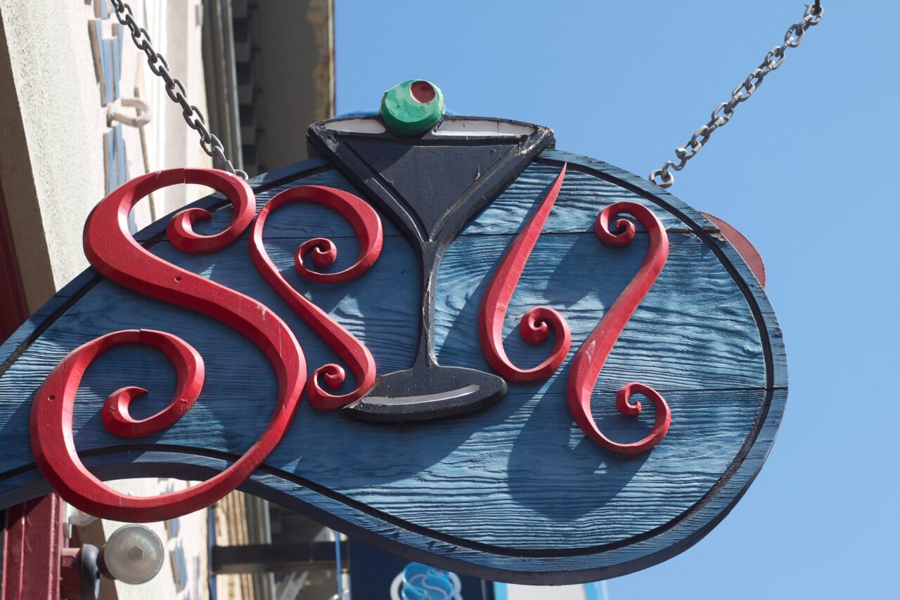 Wood store sign hanging on a chain. In the middle is a green martini like cocktail glass, and te letters around are script like letters that might read Stir.