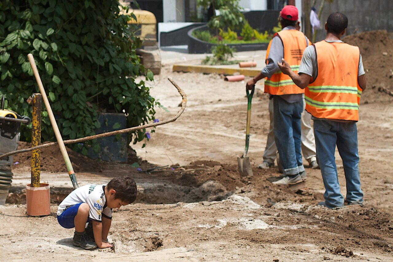 Small boy peers into hole while two construction workers look the other direction.
