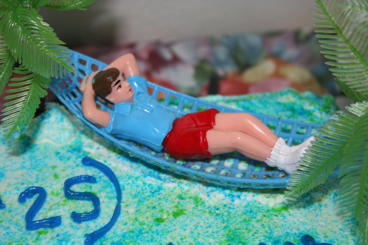 Detail of a cake dewcoration with the number 25 in lower left. A toy figure of a man in red shorts laying comfortably on a hammock suggests the dream of retirement.