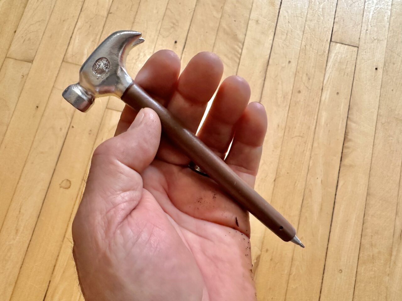 An open hand holds a pen with the top end shaped like a toy hammer. It is held over the stripes of a wood floor.