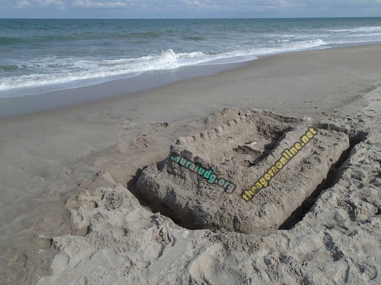 A large sand castle on a beach, with a moat around it. Superimposed on the side is muraludg.org and on the back theagoraonline.net