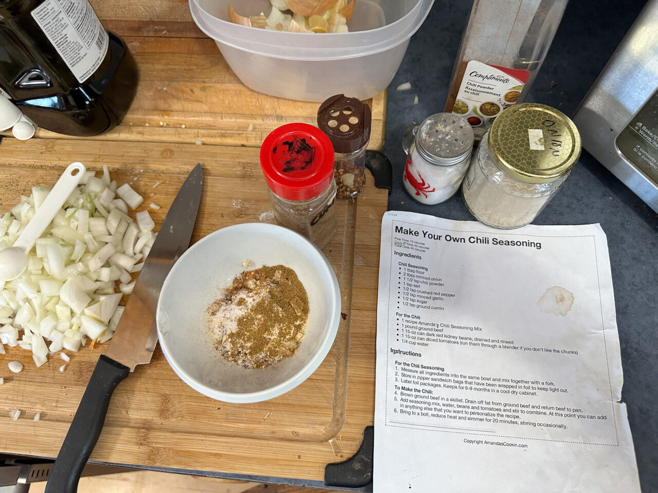 Messy kitchen counter with a knife and chopped onions on the cutting board, various spice containers and a ix of them in a bowl in the center. To the right is a printed recipe for Make Your Own Chili Seasoning. No measuring spoons are visible.