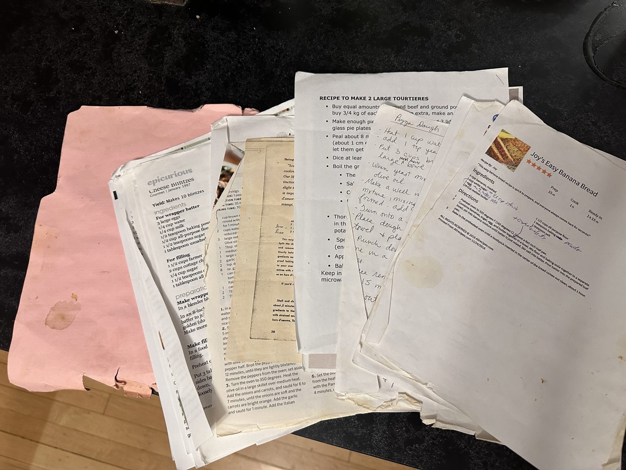 A pile of many printed recipes on worn paper spread out atop a worn pink folder sitting on a black counter top.