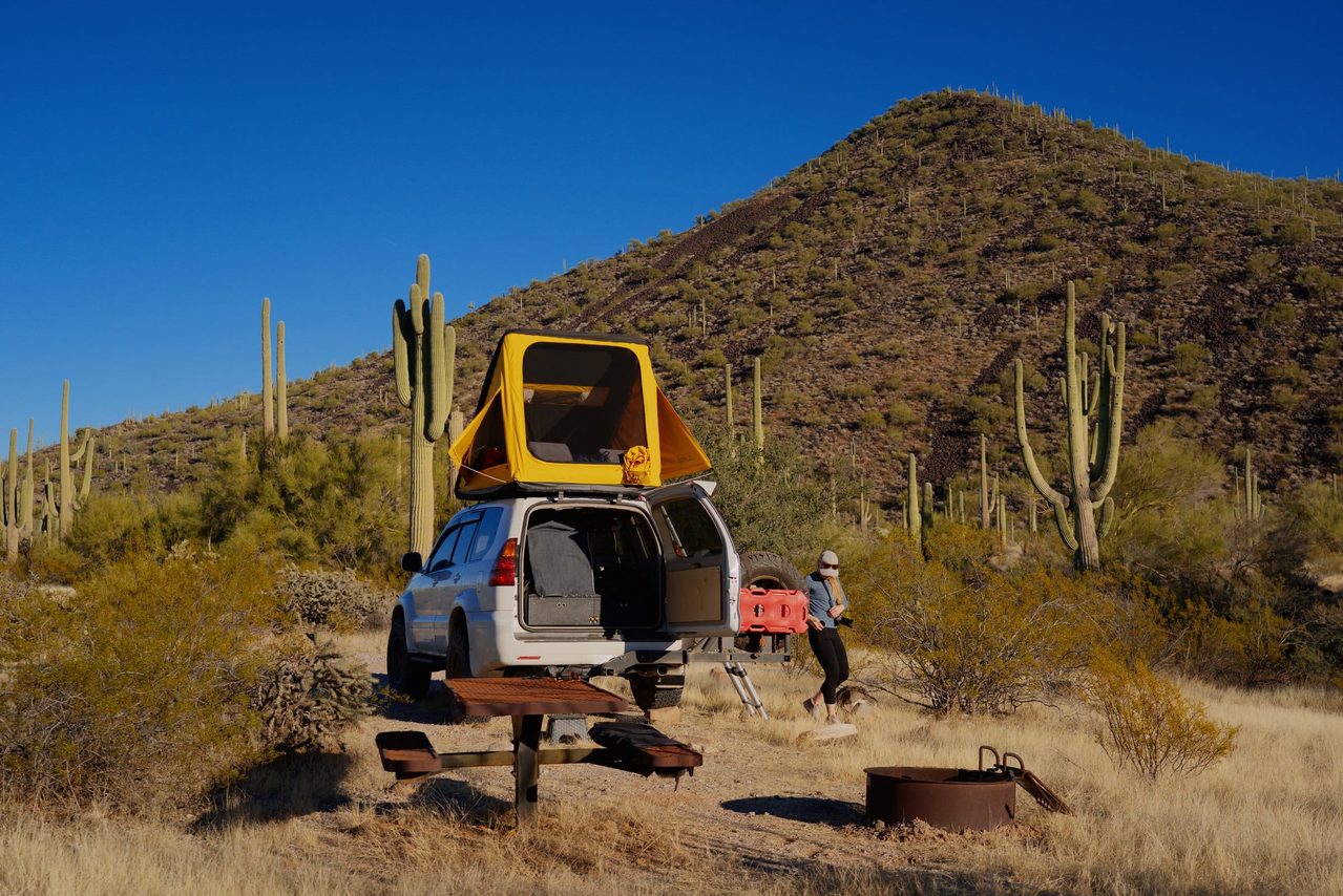A rooftop camper on an SUV parke din Arizona desert near a rusty table and a fire pit.